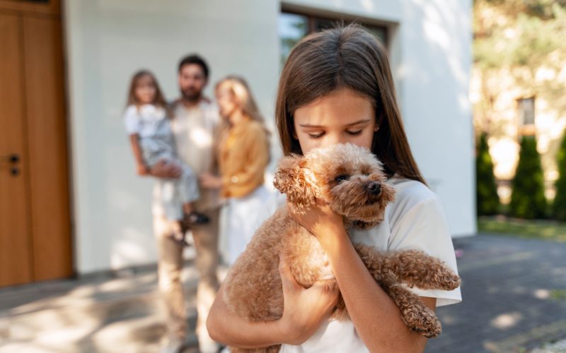 Mädchen hält Hund im Arm, im Hintergrund Familie vor neuem Haus nach erfolgreicher Baufinanzierung
